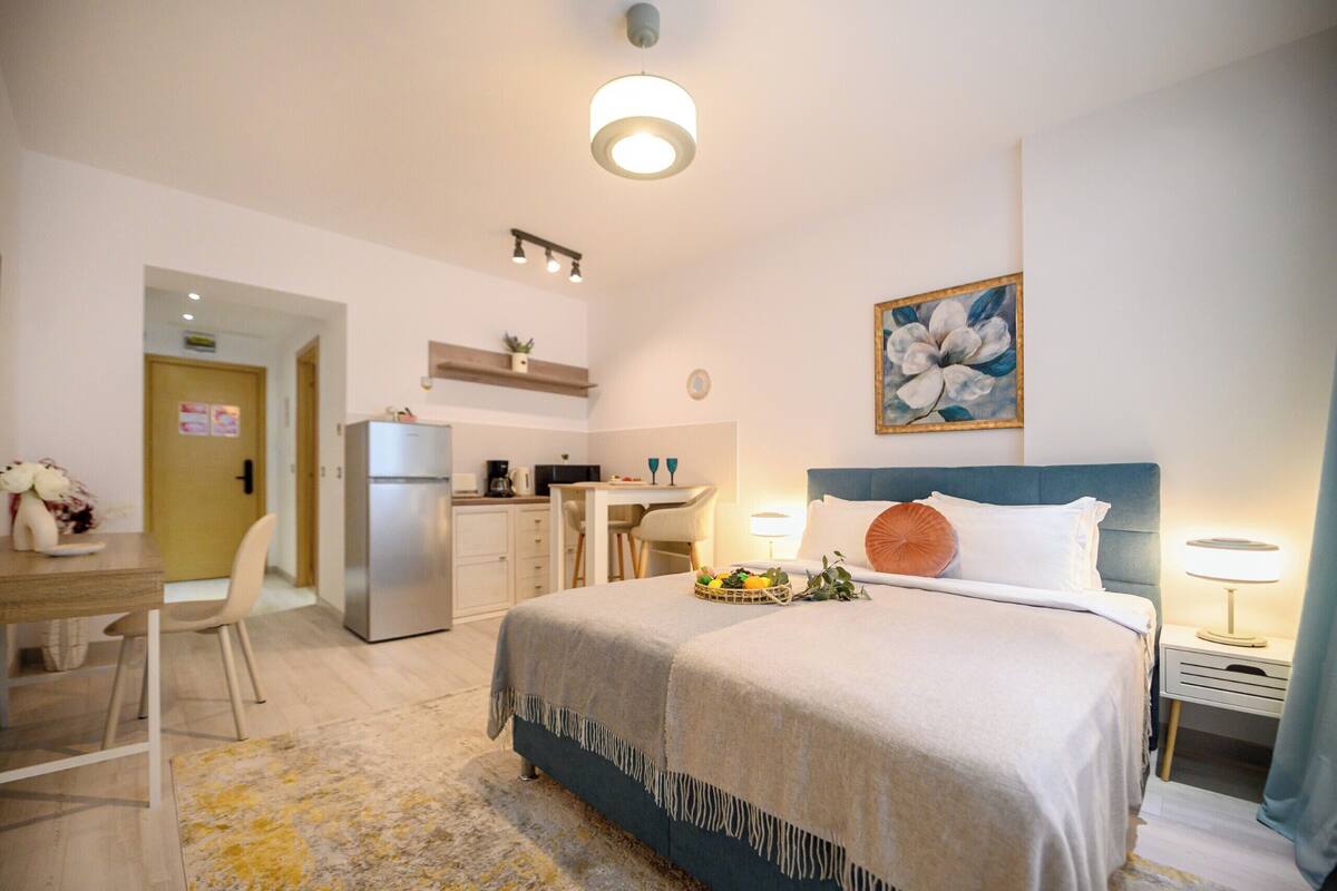 A hotel room with a bed, desk, kitchenette, and a painting on the wall.
