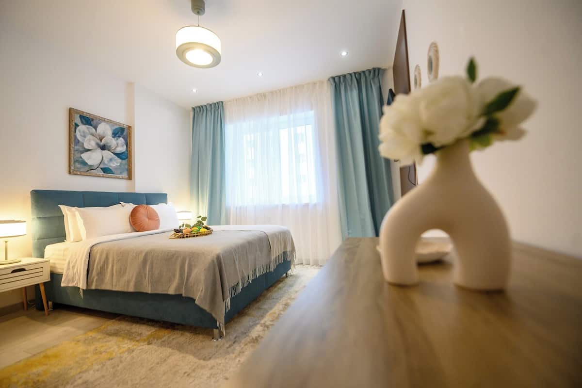 A bedroom with a bed, a nightstand, a lamp, a vase with flowers, and a painting on the wall.
