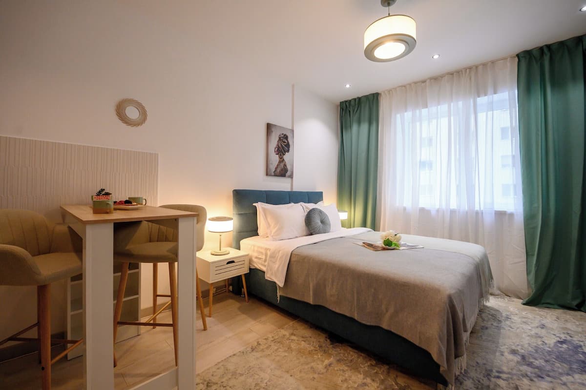 A bedroom with a bed, a desk, a chair, a nightstand, a lamp, and a window with curtains.