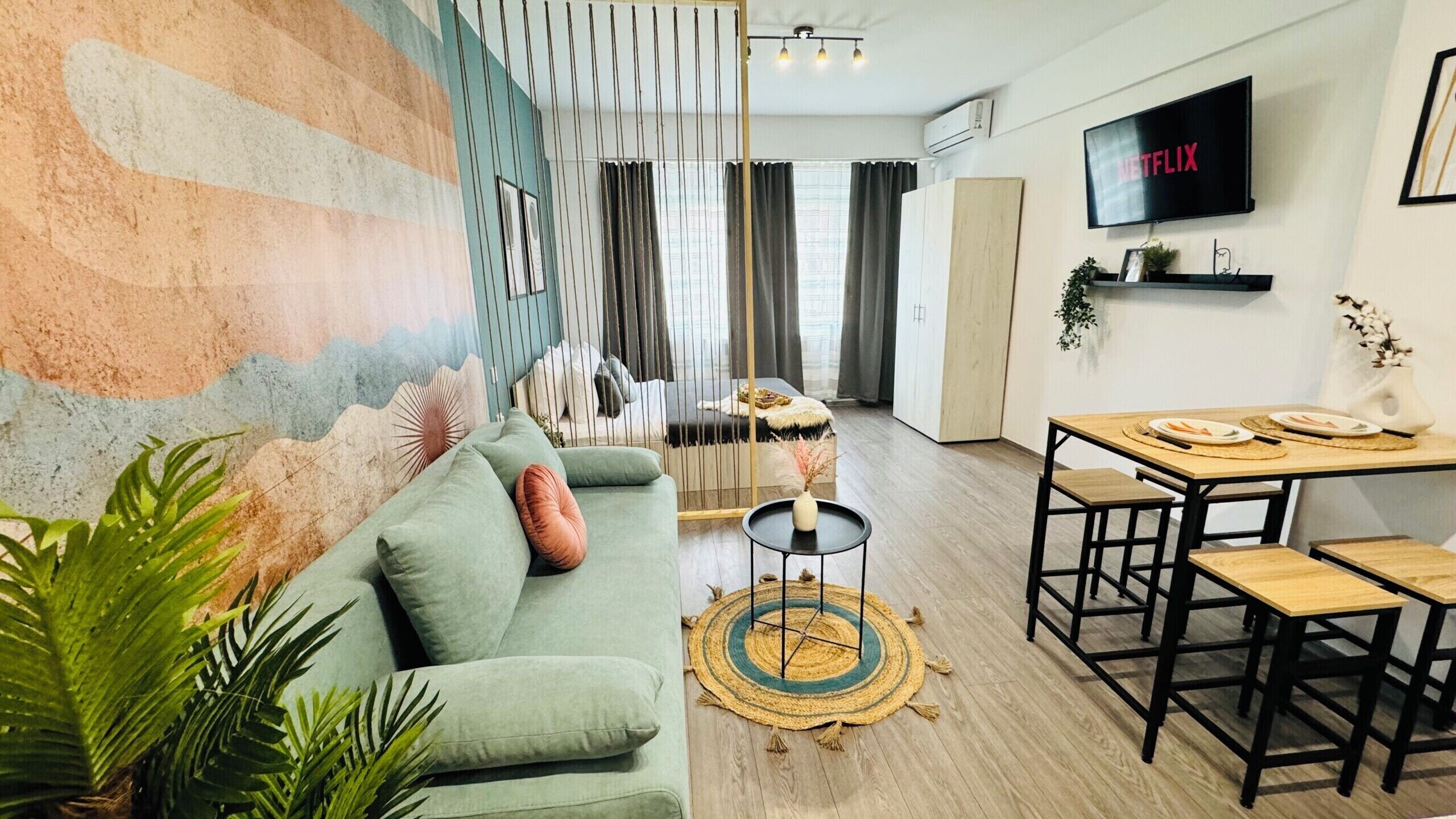 A modern living room with a sofa, a dining area with a table and chairs, a TV mounted on the wall, and a decorative wall mural.