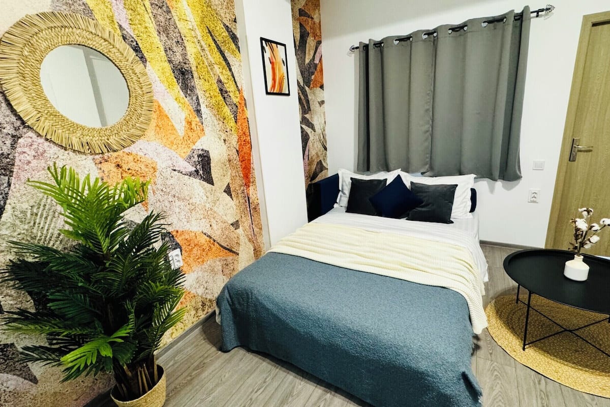 A bedroom with a bed, a round mirror, a potted plant, a small table with a vase, and a door.