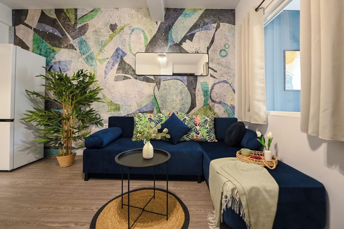A modern living room with a blue sofa, a round coffee table, a large potted plant, and a colorful abstract wall mural.