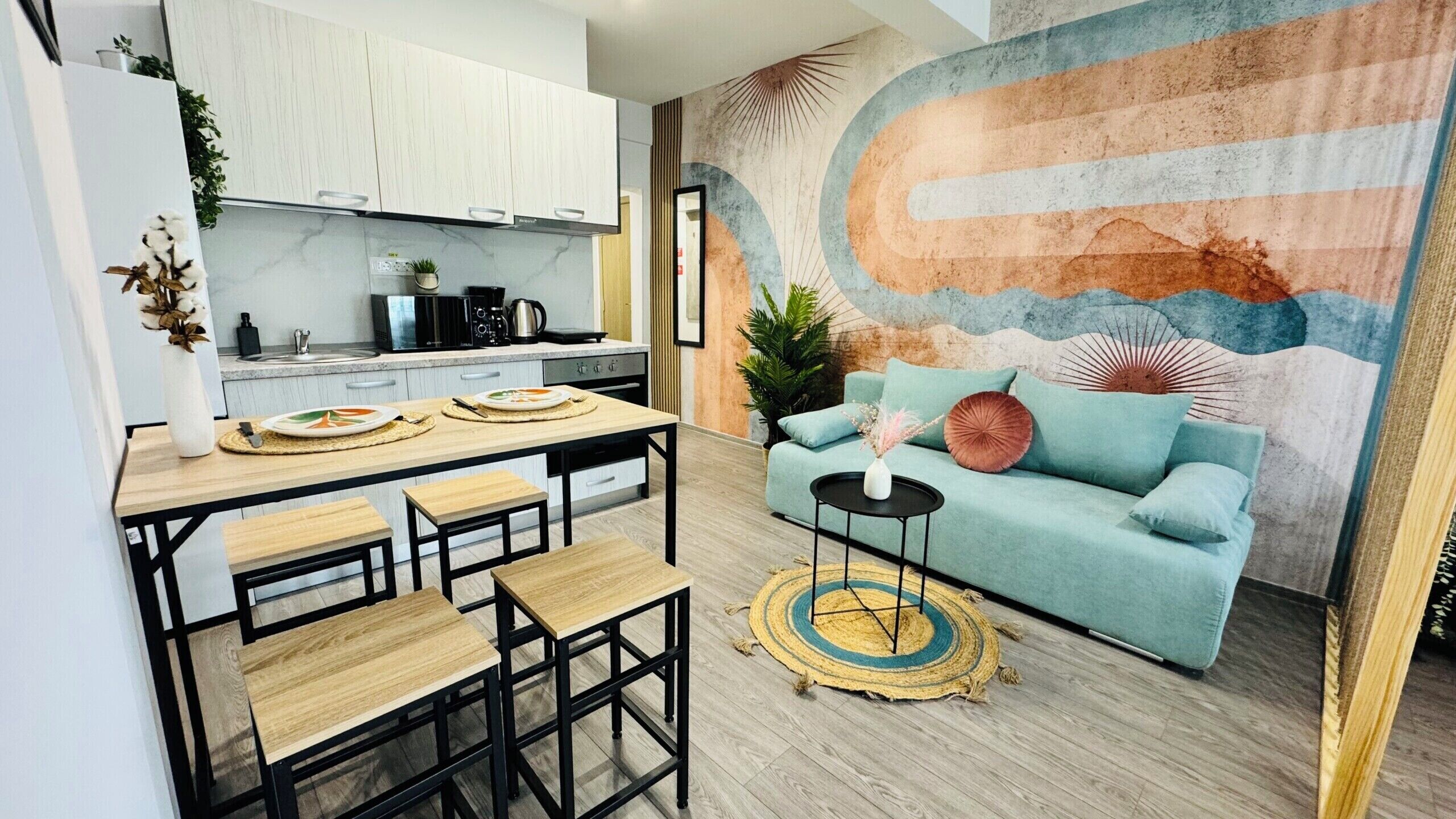 A modern kitchen with white cabinets, a dining area with a wooden table and benches, and a living room with a teal sofa and a decorative rug.