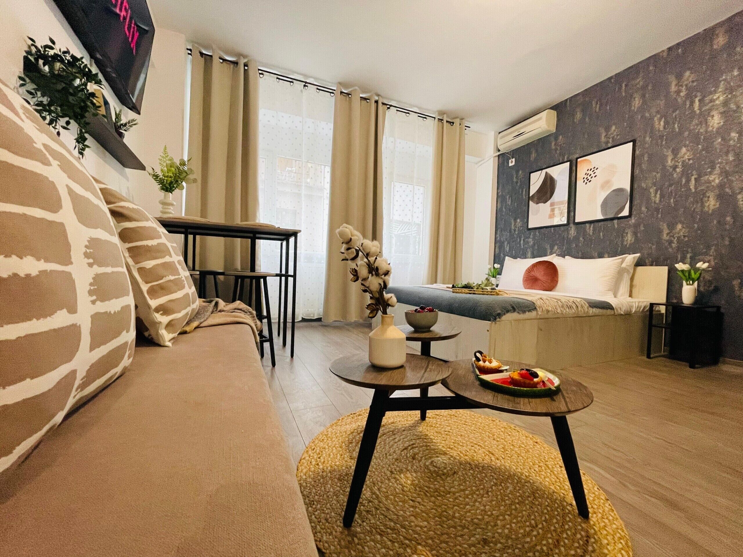 A modern living room with a sofa, a dining table, a small round table with a bowl and fruit, a bed with a blue headboard, and a wall-mounted air conditioner.