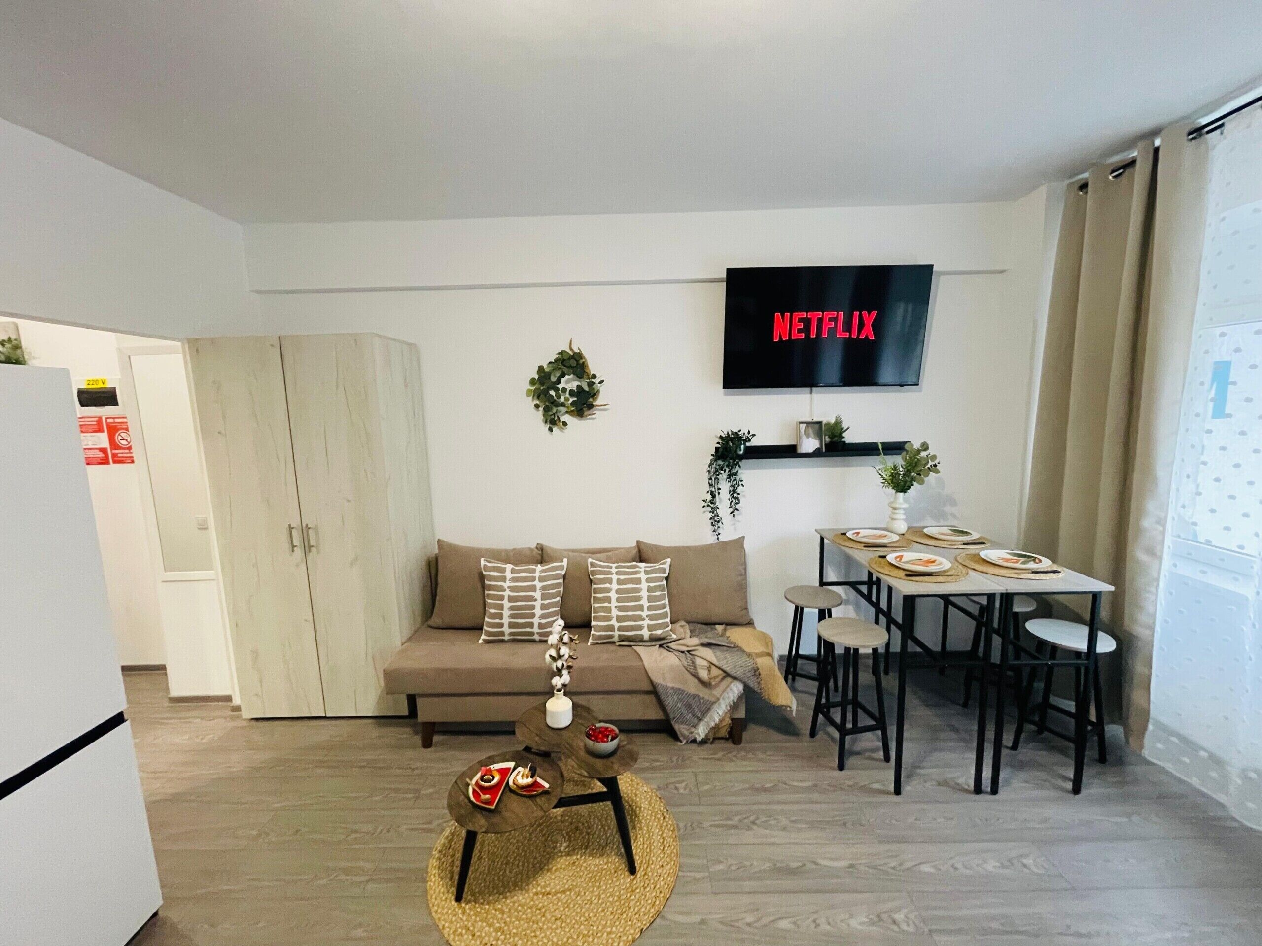 A modern living room with a sofa, coffee table, dining table, and chairs. A flat-screen TV mounted on the wall displays 'NETFLIX'.