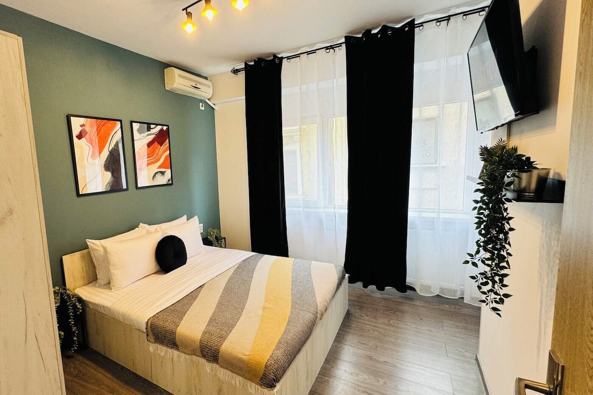 A bedroom with a bed, a wall-mounted air conditioner, a window with black curtains, and a plant.