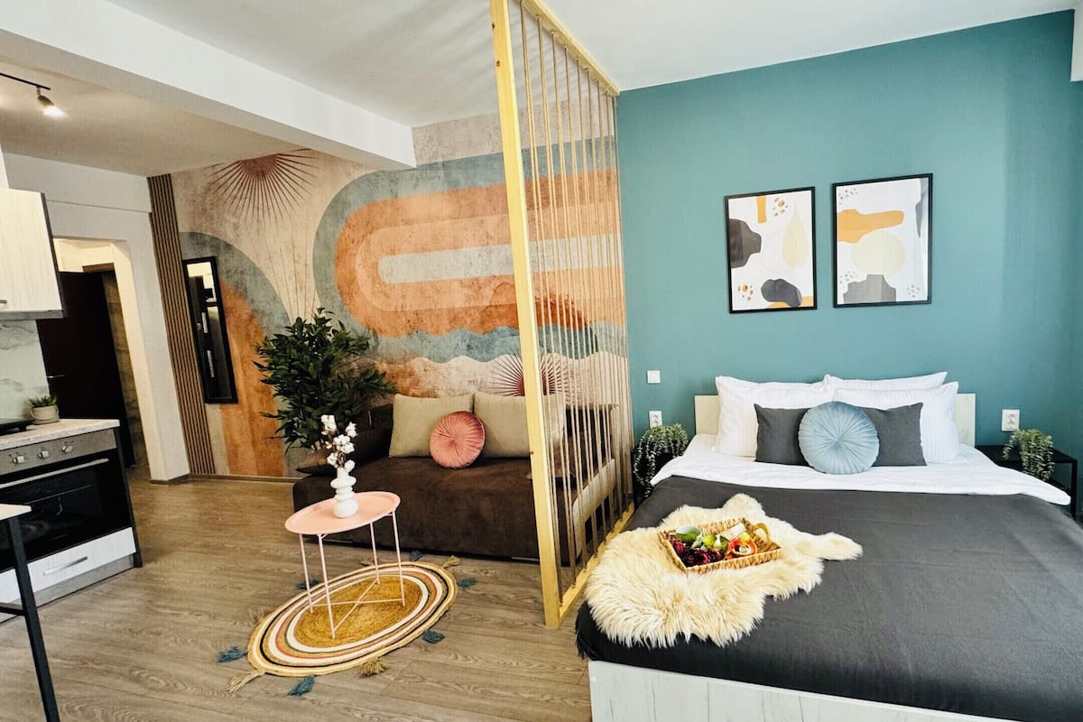 A modern bedroom with a bed, a small side table, a decorative wall piece, and a view into the kitchen.