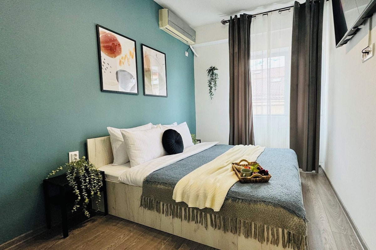 A bedroom with a bed, a window with curtains, a wall-mounted air conditioner, and two framed artworks.