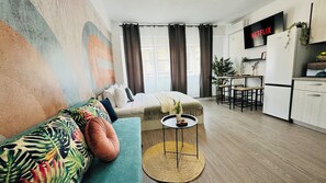Un apartamento moderno con sofá, una pequeña mesa redonda, zona de cocina y un amplio ventanal con cortinas.