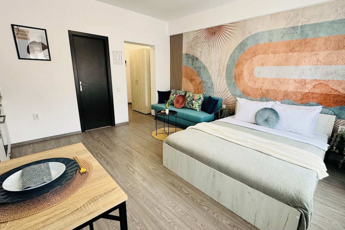 A modern hotel room with a bed, a small table, a sofa, and a large abstract wall mural.