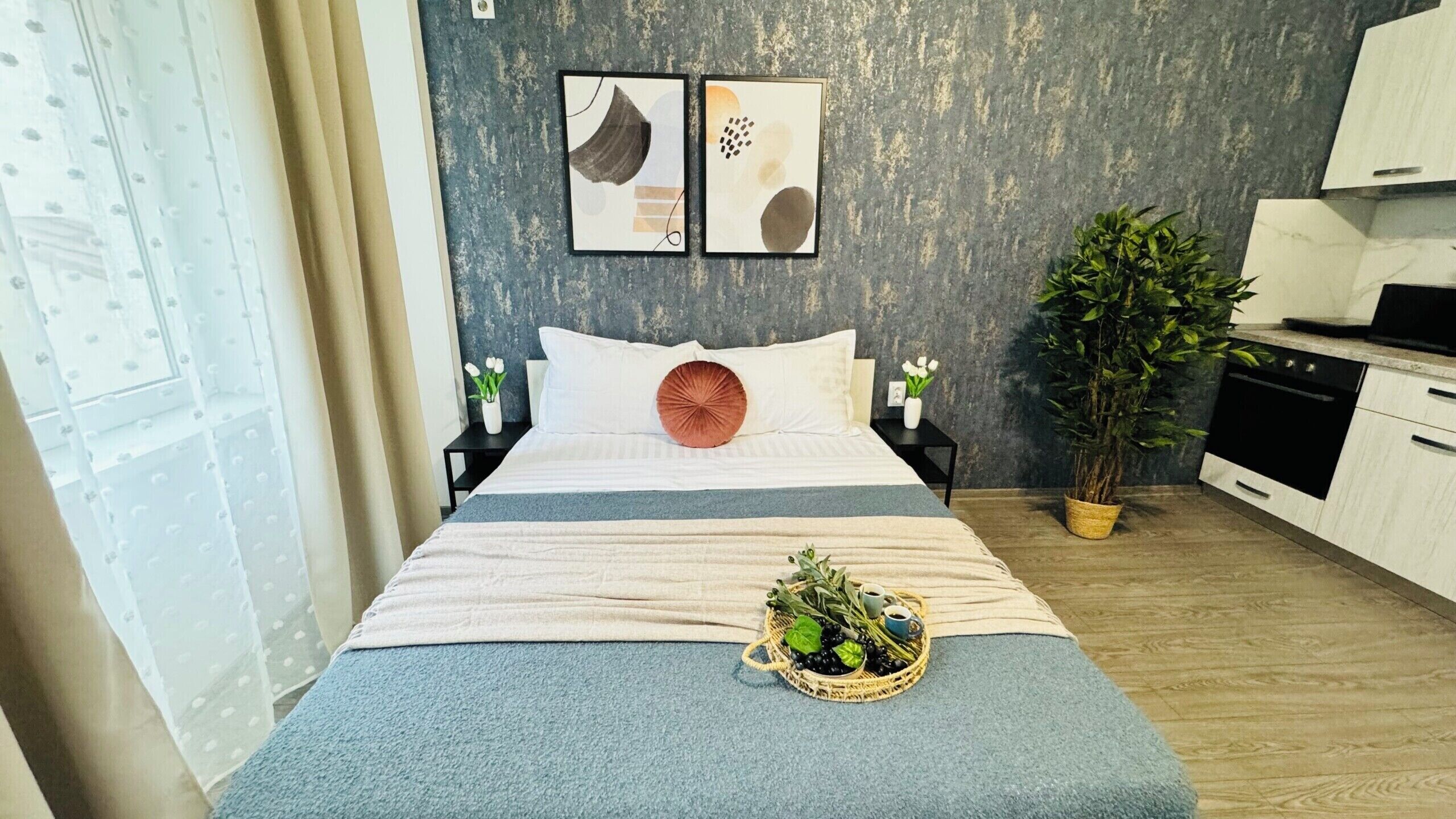 Habitación de hotel con cama, una mesita con una planta, una zona de cocina y obras de arte en la pared.