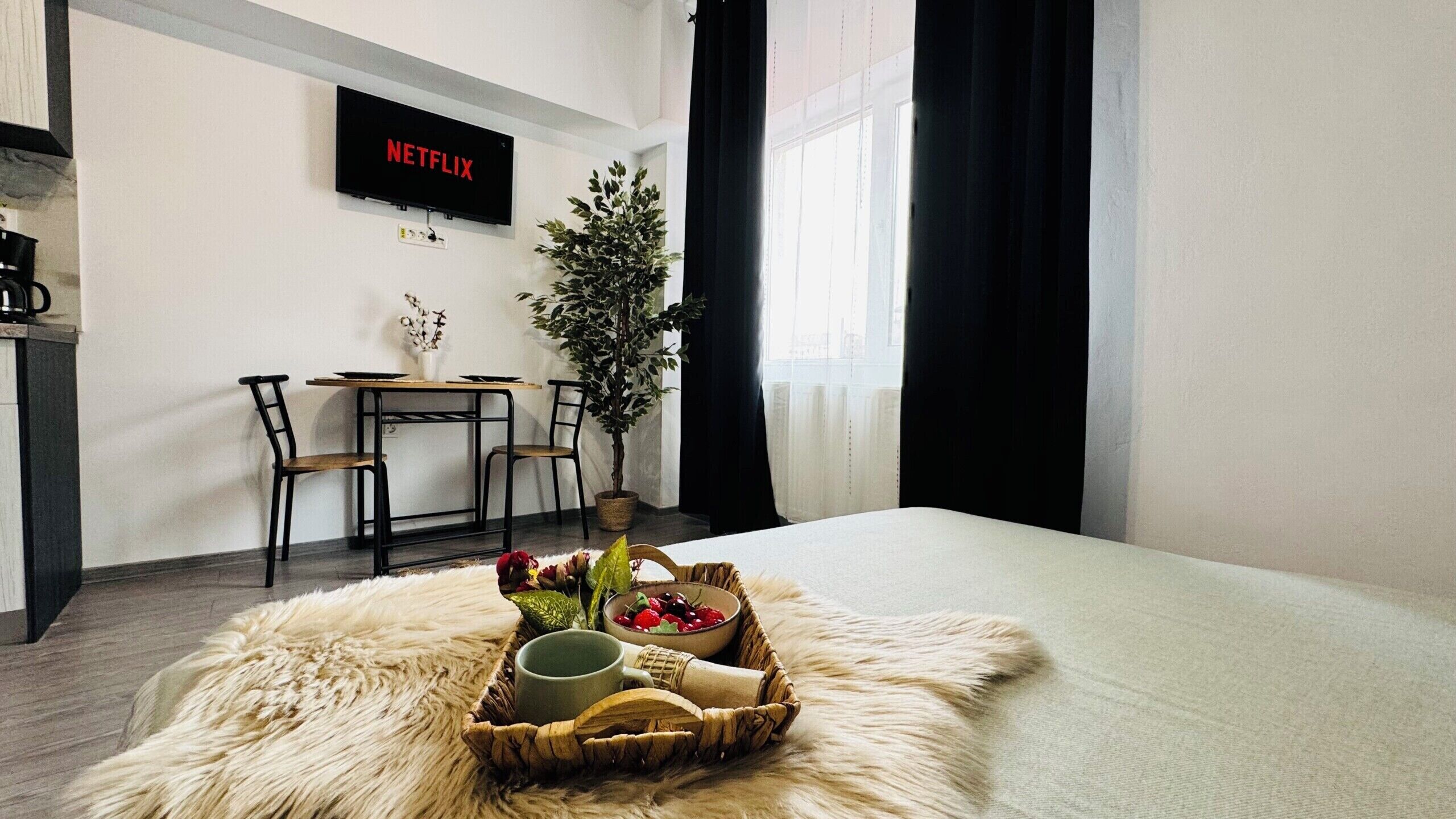 A hotel room with a bed, a dining table with chairs, a television displaying 'NETFLIX', a potted plant, and a basket with fruits and a bottle.