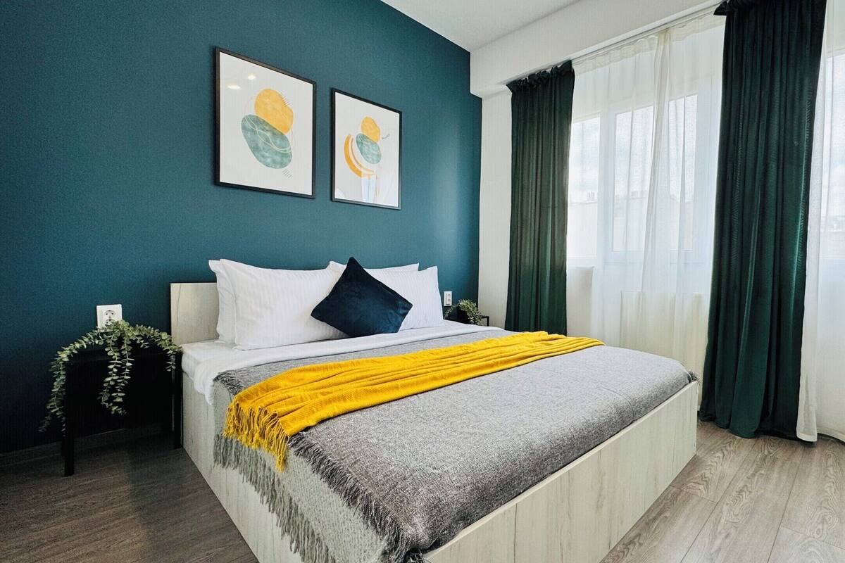 A bedroom with a bed, a nightstand, a window with curtains, and two framed artworks on the wall.