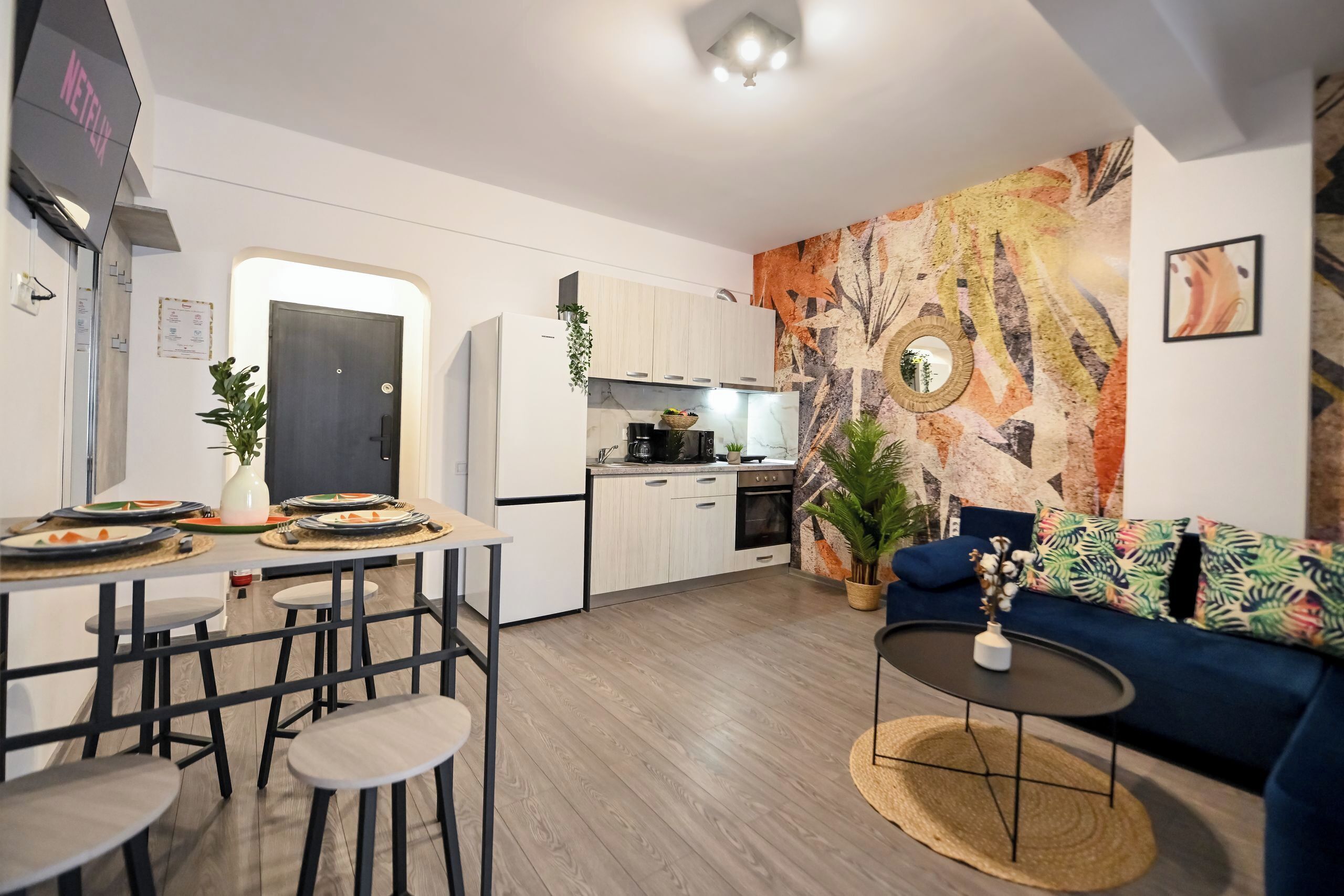 Cozinha moderna com armários brancos, área de refeições com banquetas, sofá azul e parede com arte abstrata.
