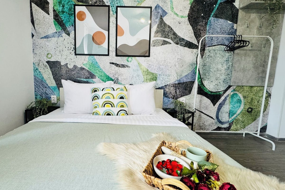 A modern bedroom with a bed, pillows, a wall art piece, and a bedside table with a plant.