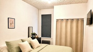 A bedroom with a bed, a bedside table, a mirror, a window with blinds, and a TV mounted on the wall. - FIFA World Cup Stay • Easy Transit to MetLife • Near NYC (Jersey City)