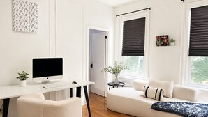 A modern living room with a white desk, computer, and chair, a sofa with a blanket, and a window with a view. - FIFA World Cup Stay • Easy Transit to MetLife • Near NYC (Jersey City)