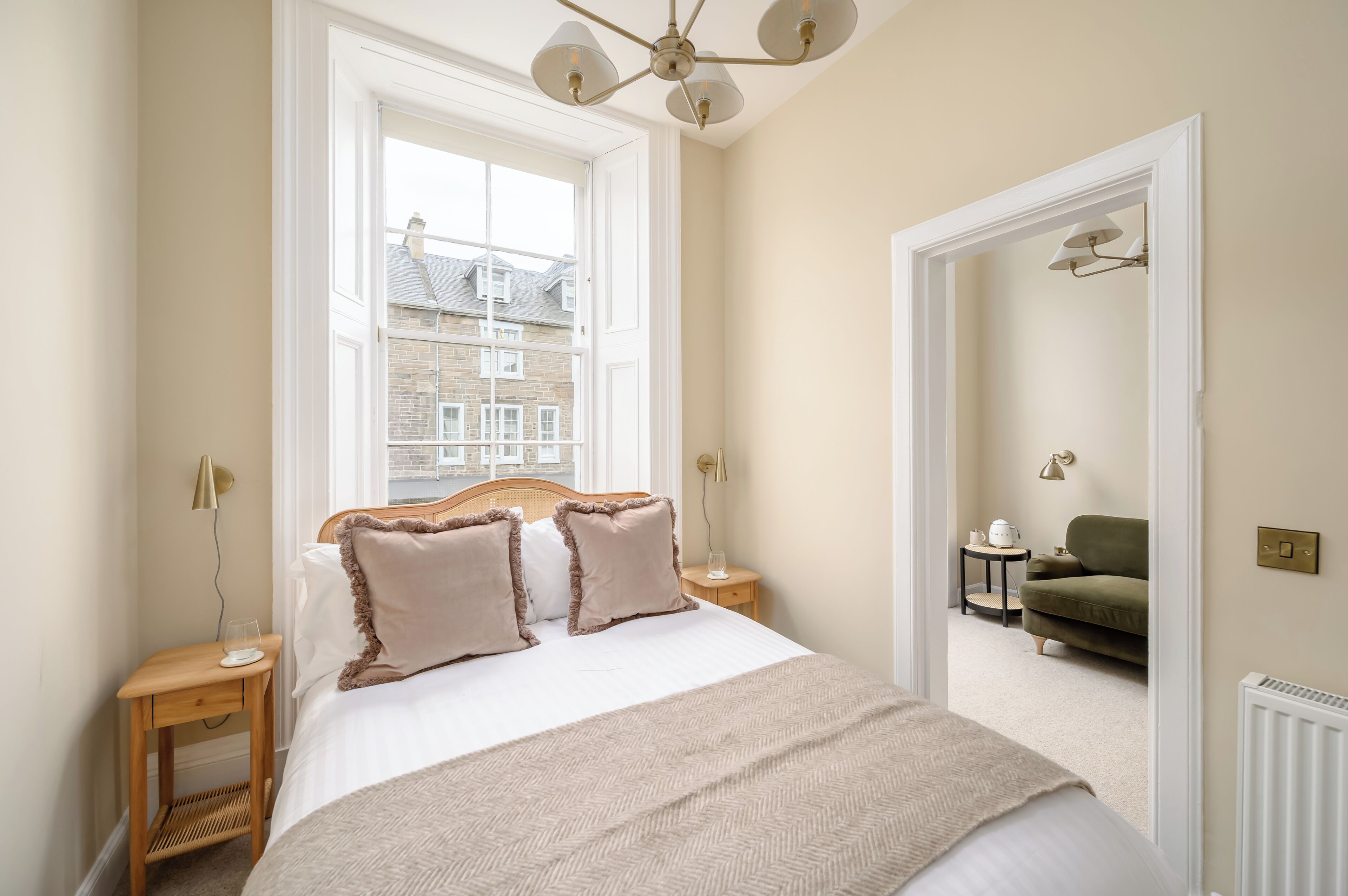 A bedroom with a large bed, a wooden nightstand, a bedside table, a window with a view of buildings, and a door leading to another room.