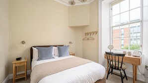 Un dormitorio con cama, mesitas de noche, escritorio, silla, ventana y radiador.