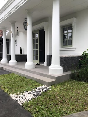 Un edificio con un porche cubierto sostenido por columnas blancas, un camino de piedra que conduce a la entrada y un pequeño jardín con césped y piedras.
