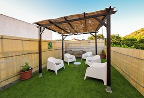 Un espacio al aire libre techado con césped artificial, sillas blancas y una pérgola con luces colgantes.