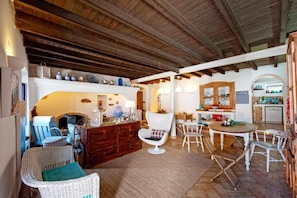 A rustic interior with a wooden ceiling, a dining area with white chairs, a kitchen with wooden cabinets, and a fireplace. - Casa da Tia Amália (Mértola)
