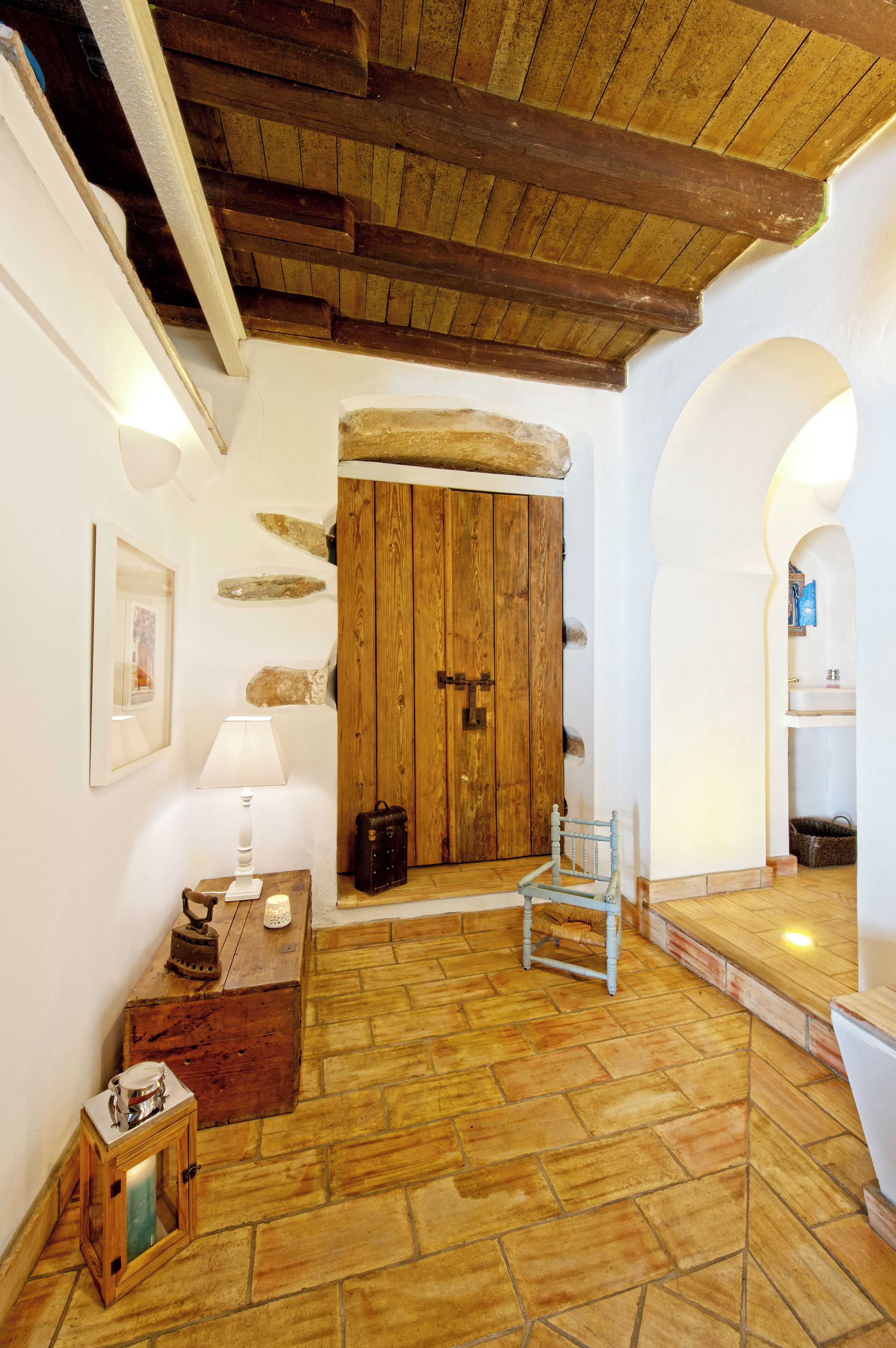 Un interior rústico con una puerta de madera, paredes de piedra, una pequeña mesa con una lámpara y una silla.