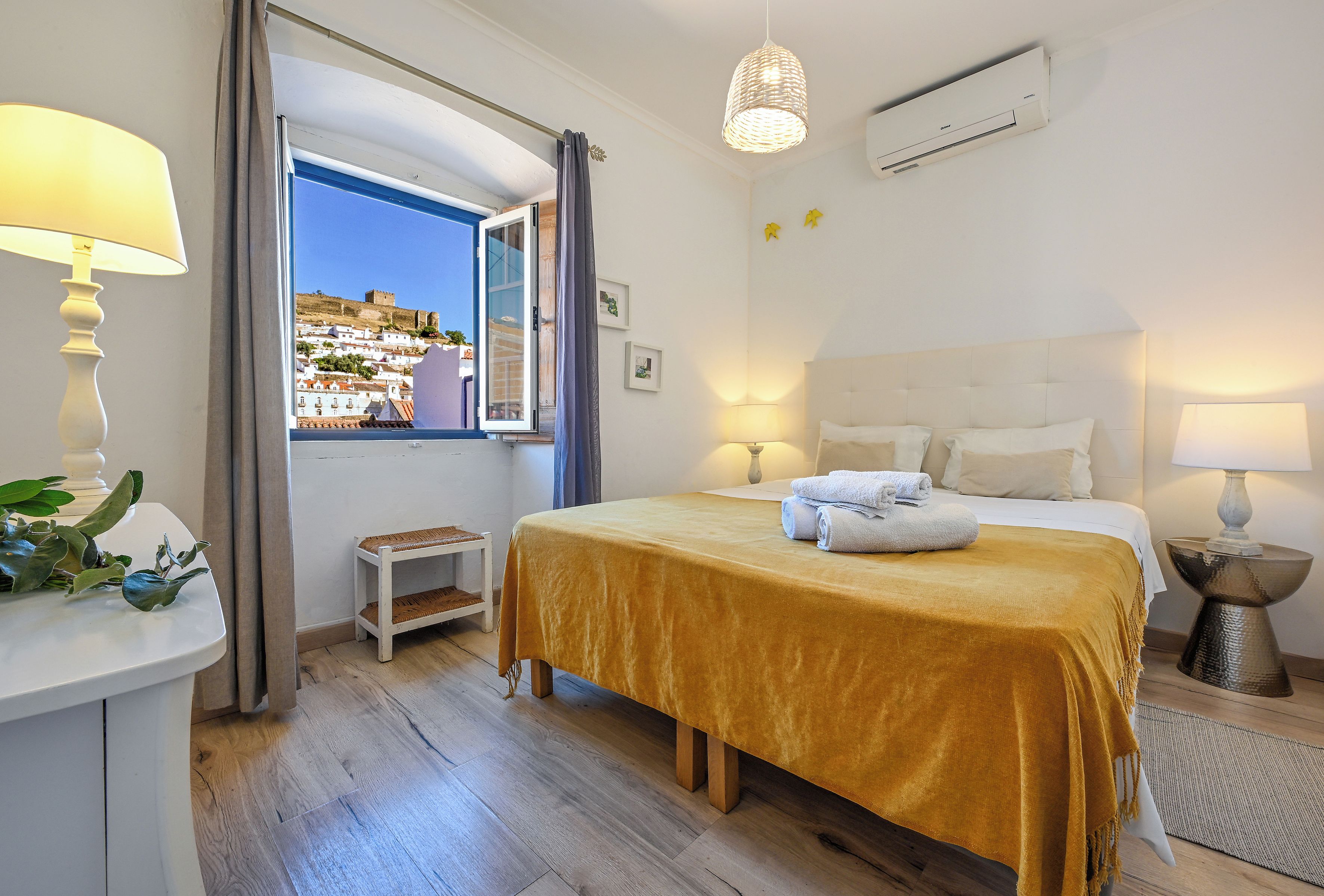 A hotel room with a bed, bedside lamps, a window with curtains, a view of a hill, and an air conditioning unit.