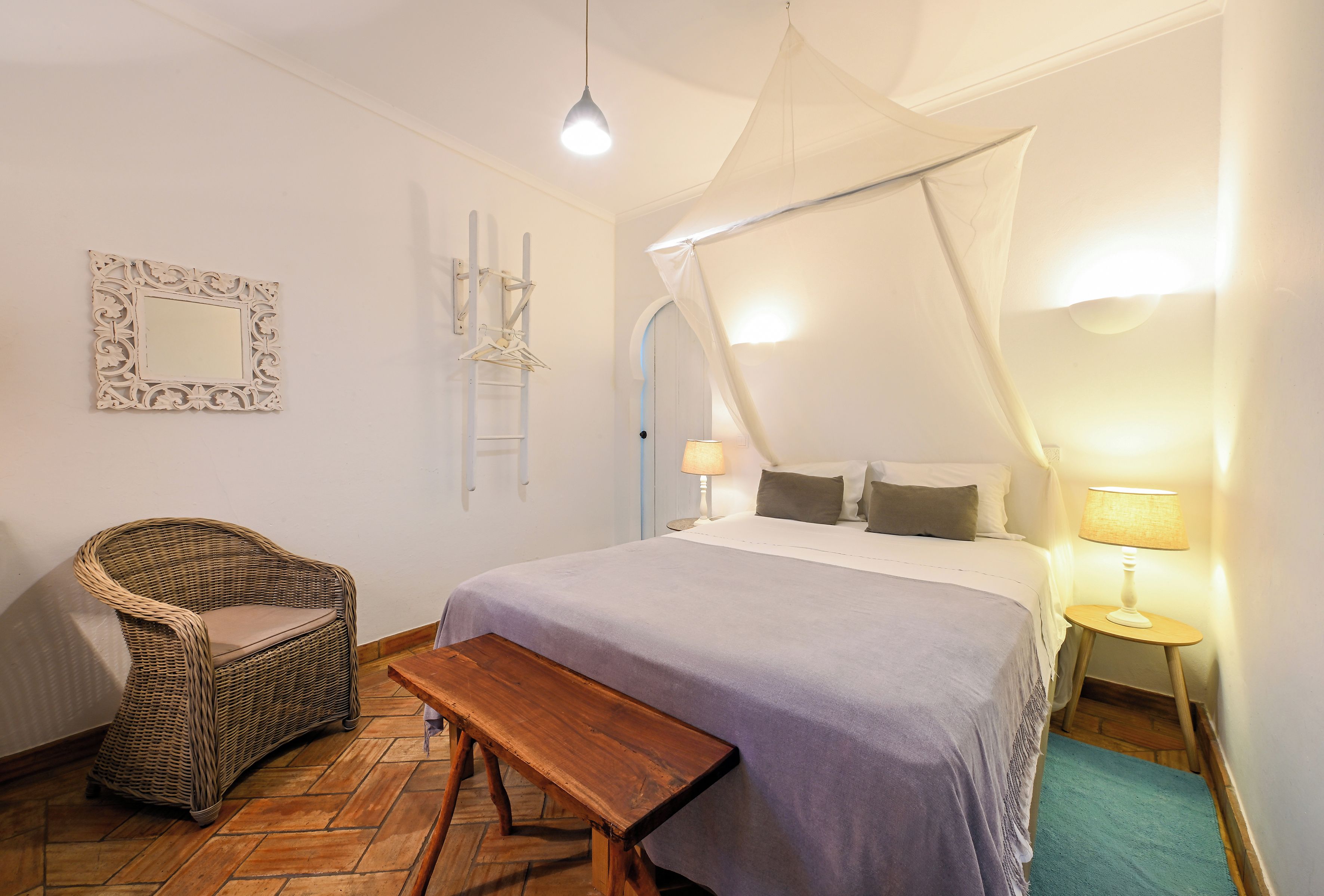 A bedroom with a bed, a bench, a chair, a hanging light fixture, and a wall-mounted mirror.