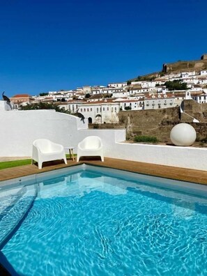 A swimming pool with two white chairs, a large white sphere, and a view of a hillside with buildings.