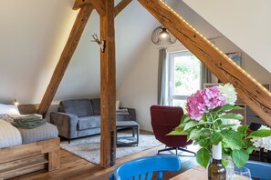 Quarto em estilo loft com teto de madeira, uma cama, um sofá cinzento, uma poltrona vermelha e um vaso com flores.