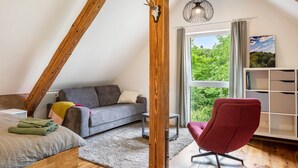 Un dormitorio tipo loft con techo inclinado, una cama, un sofá gris, un sillón rojo, una estantería de madera y una ventana con vistas a zonas verdes.