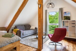 Un dormitorio tipo loft con techo inclinado, una cama, un sofá gris, un sillón rojo, una estantería de madera y una ventana con vistas a zonas verdes.