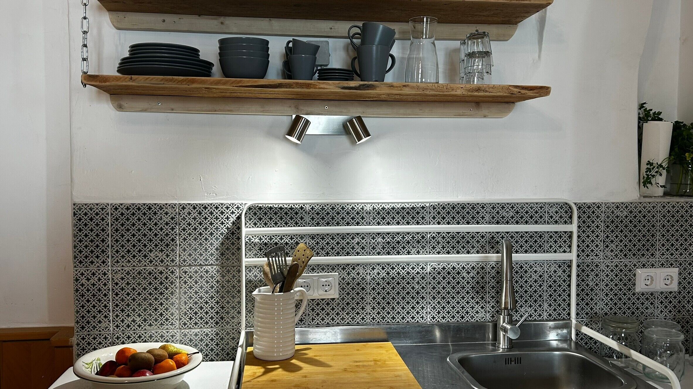 Eine Küche mit Holzregal, Spüle, Obstschale und backsplash-Fliesen mit Muster.