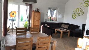 A living room with a sofa, a dining table with chairs, a lamp, and a TV. - Holiday Home with Hot Tub and Sauna near Bernsteinsee (Wiefelstede)