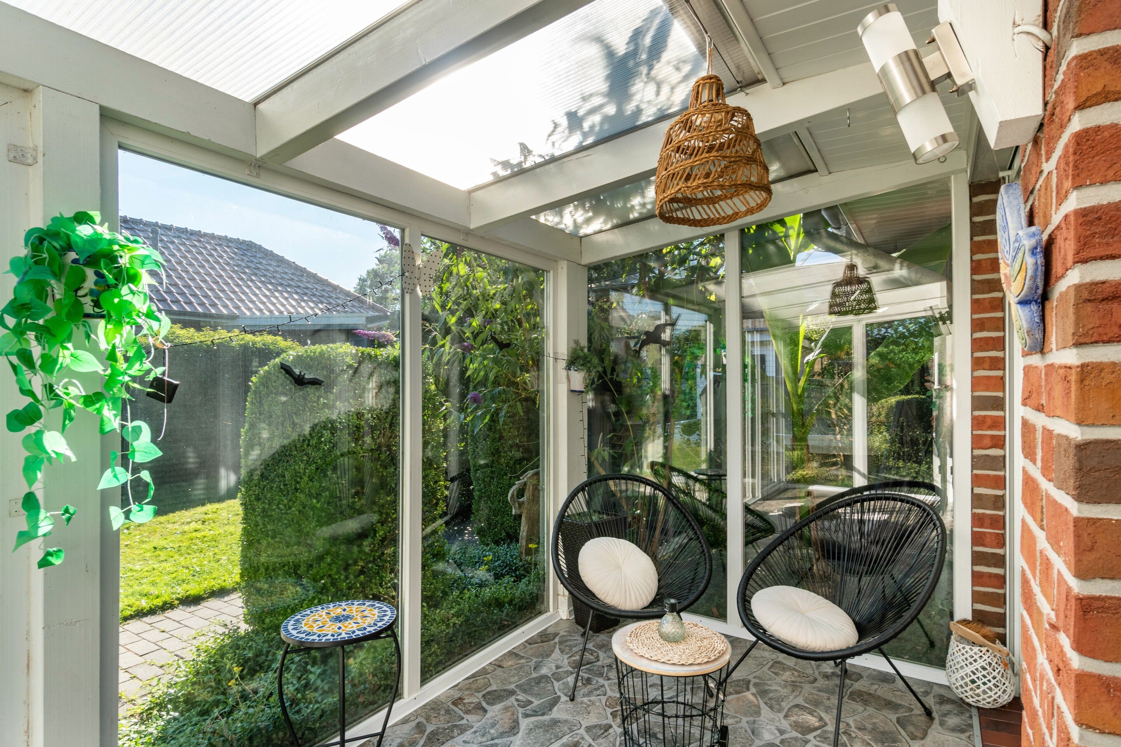 Een oranjerie met glazen deuren, rieten stoelen, een klein tafeltje en een hanglamp.