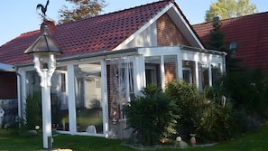 A conservatory with a glass roof, surrounded by greenery and a brick house in the background.