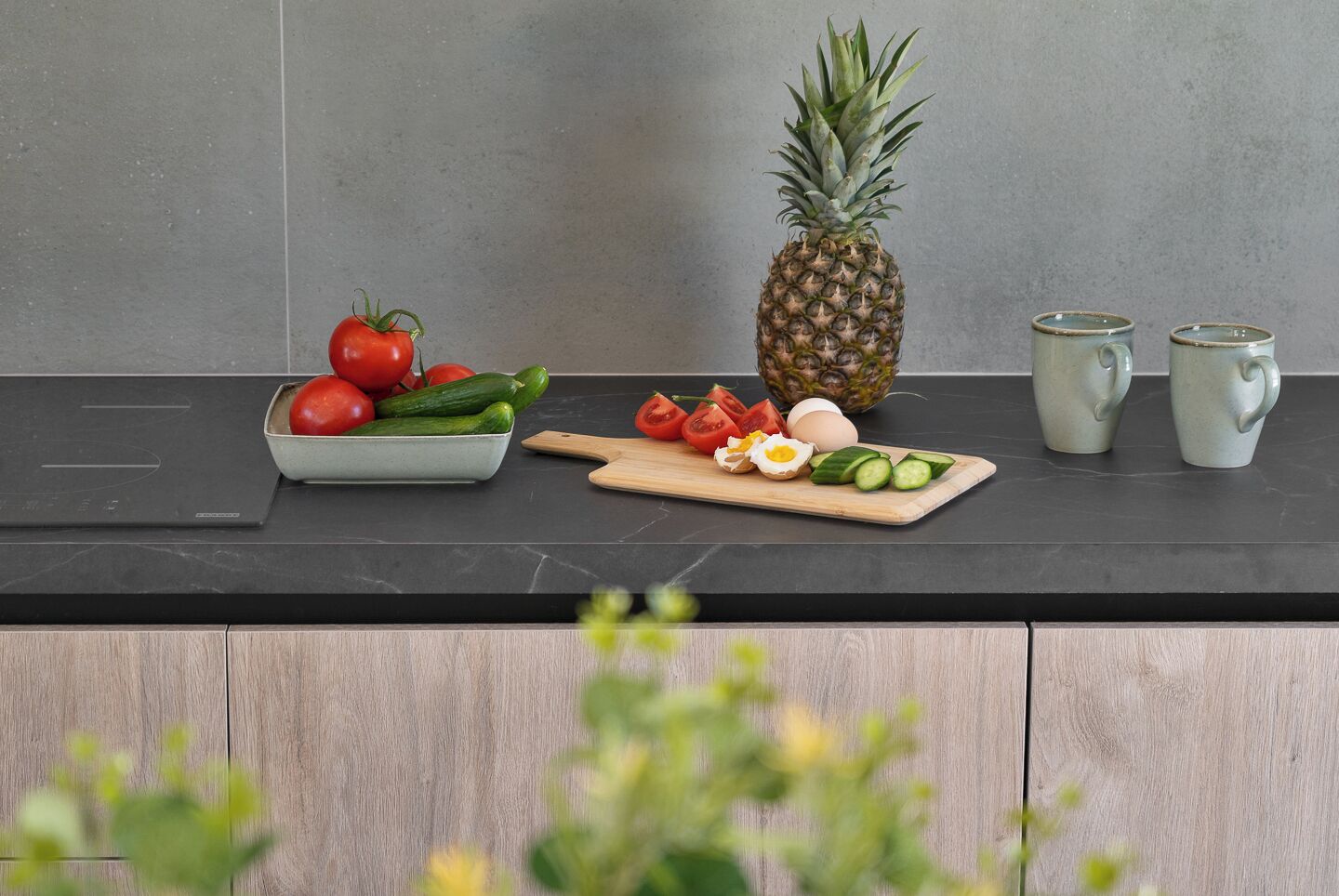 Una encimera de cocina con un tazón de tomates, pepinos y una piña, una tabla de cortar con verduras en rodajas, y dos tazas.