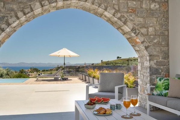 An outdoor seating area with a stone archway, a pool, and a view of the landscape. - Villa Meraki - Sea View (Chania)