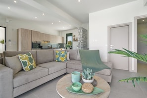 A modern living room with a grey sofa, a round coffee table, and a kitchen in the background. - Villa Meraki - Sea View (Chania)