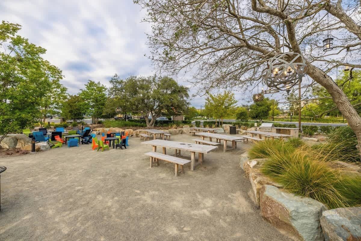 An outdoor seating area with tables and chairs, surrounded by trees and a decorative metal sculpture.