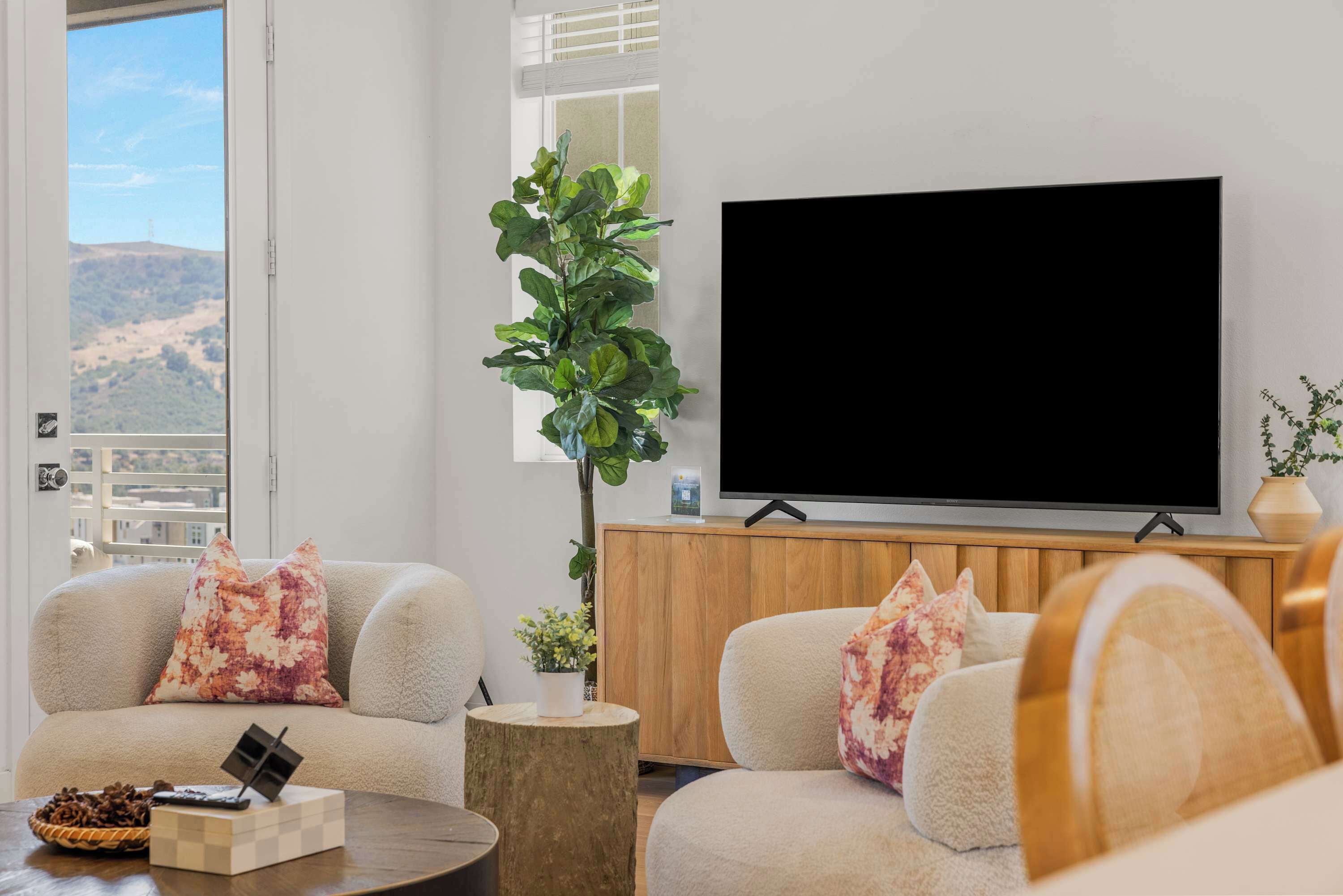 Una sala de estar con un televisor de pantalla plana grande, dos sofás con cojines estampados, un mueble de madera, una planta en maceta y vistas a una colina a través de una ventana.