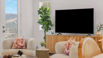 A living room with a large flat-screen TV, two sofas with floral cushions, a wooden cabinet, a potted plant, and a view of a hill through a window.