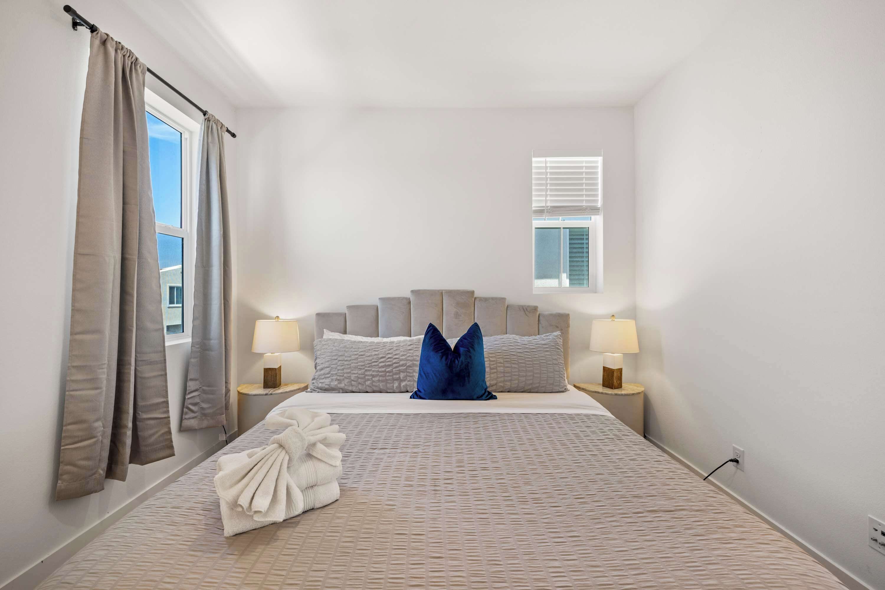 A bedroom with a bed, two bedside lamps, a window with blinds, and a towel on the floor.
