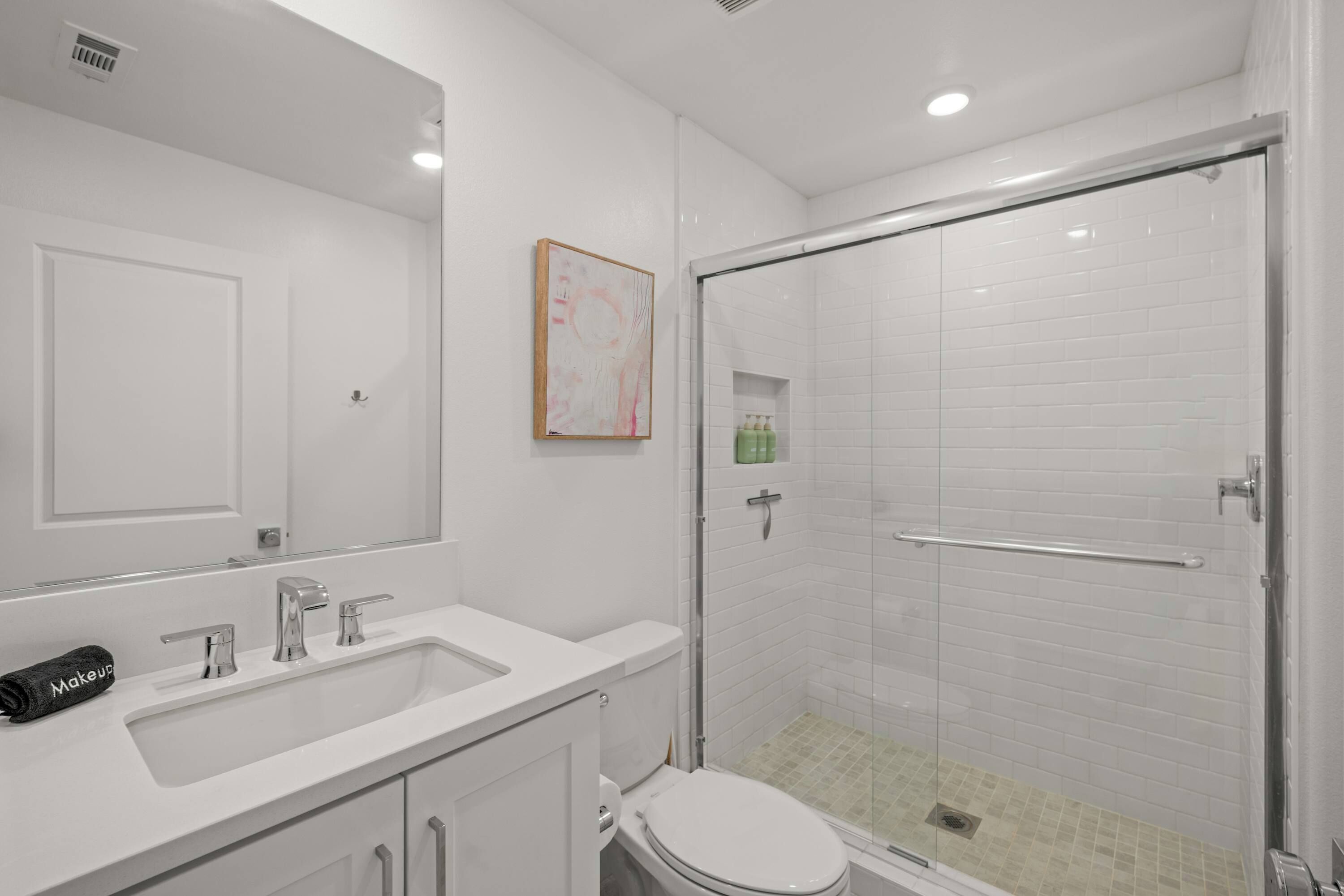 Un baño con lavamanos blanco, espejo, inodoro y cabina de ducha de vidrio, además de un cuadro en la pared.