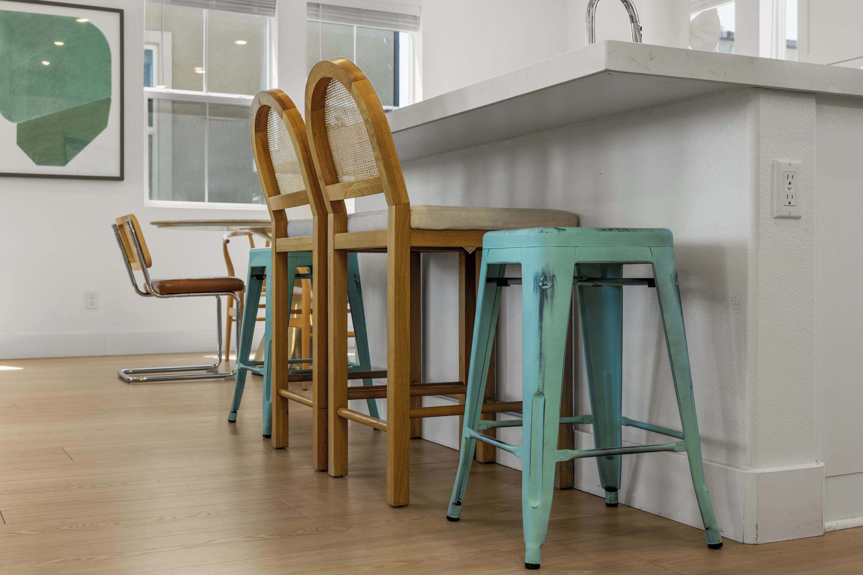 A kitchen with wooden chairs and teal stools, a window, and a white countertop.