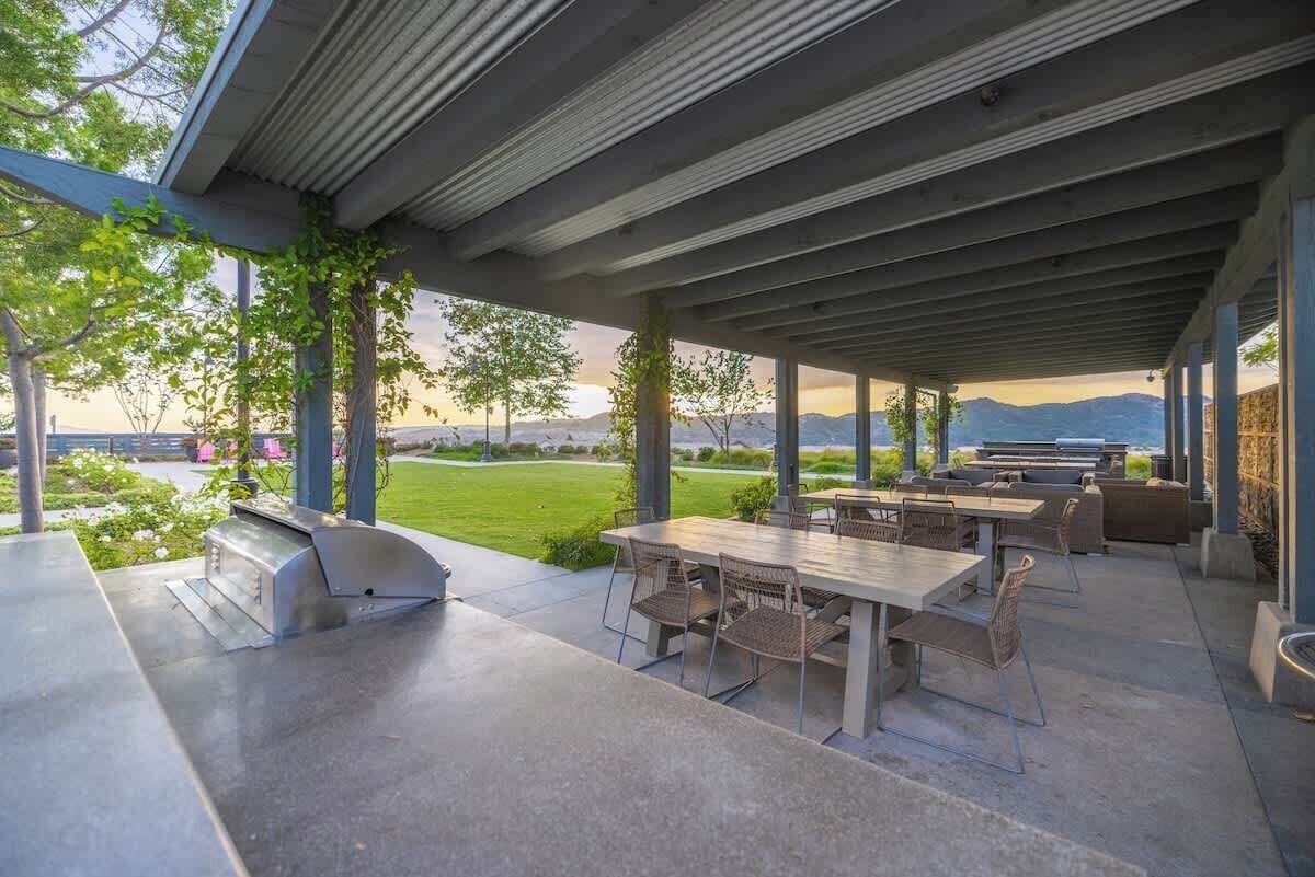 En udendørs terrasse med grill, spisebord og stole, med udsigt over en anlagt have og fjerne bjerge.