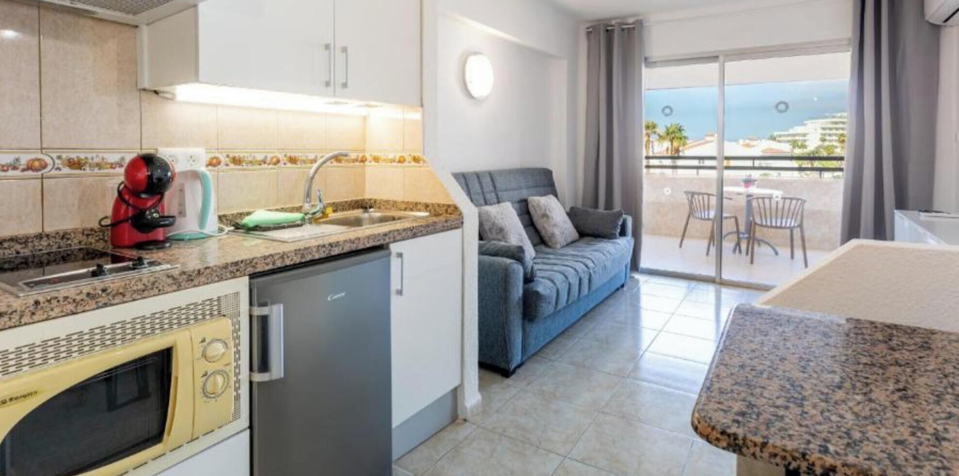 Una cucina moderna con lavastoviglie, bollitore rosso e microonde. C'è un divano grigio e un'area pranzo con vista mare.