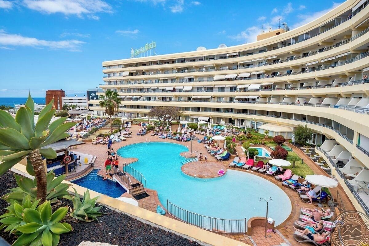 A multi-story hotel with a large swimming pool, sun loungers, and a view of the sea.