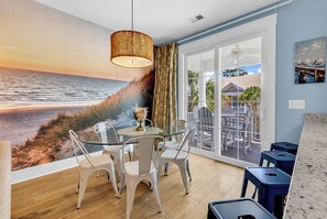 Ein Zimmer mit Meeresblick, an der Wand ein Strandgemälde, ein Glastisch mit Stühlen und ein Zugang zu einer Außenbereich-Sitzgelegenheit durch eine Glastür.
